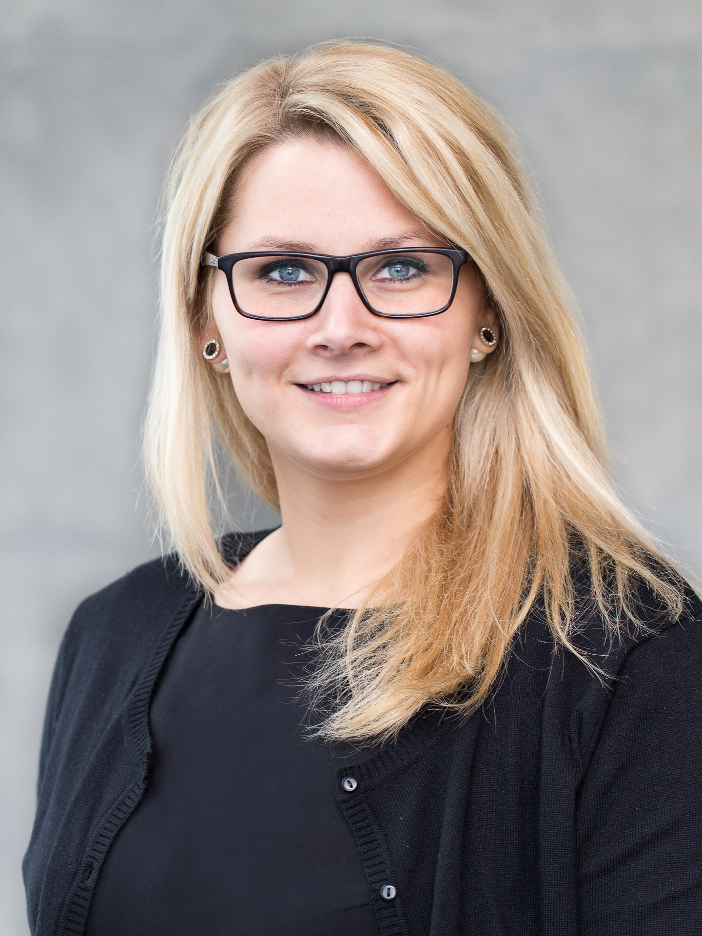 Portraitfotografie - Business Portraits: Junge Frau in schwarzer Kleidung und mit Brille stehend vor grauer Wand