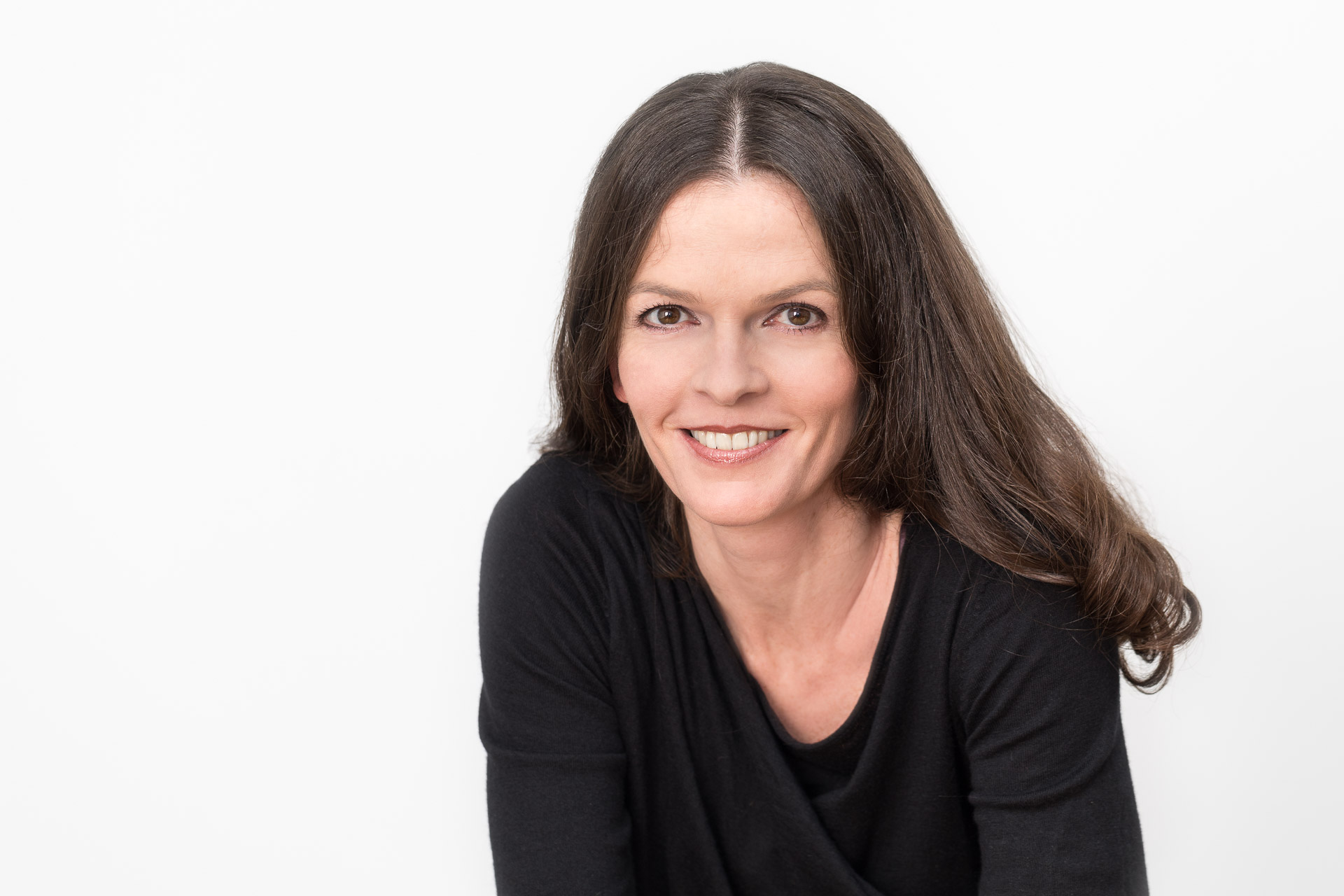 Portraitfotografie - Business Portraits: Lächelnde Geschäftsfrau, schwarz gekleidet, stehend vor weißer Wand.