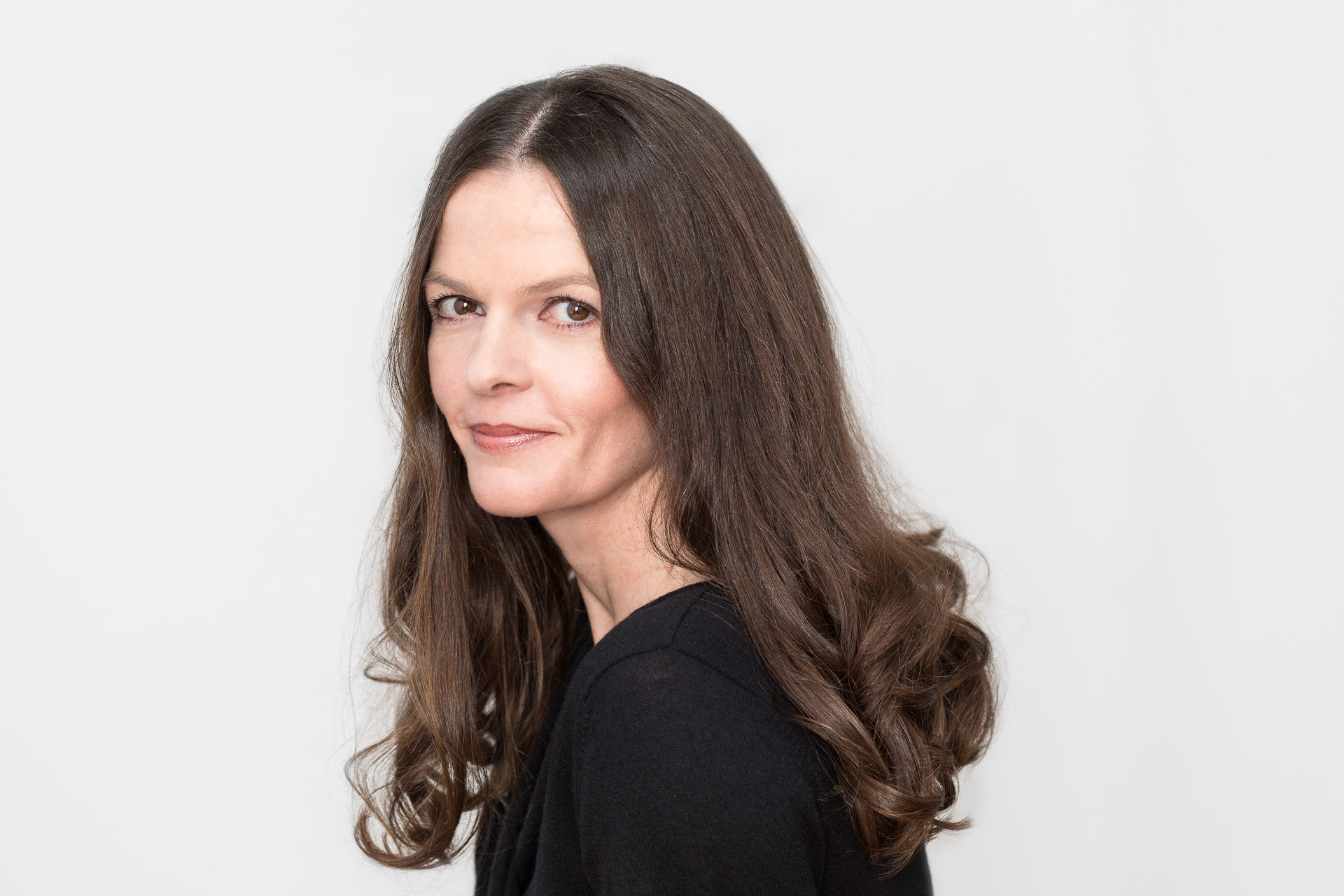 Portraitfotografie - Business Portraits: Lächelnde Geschäftsfrau, schwarz gekleidet, stehend vor weißer Wand.