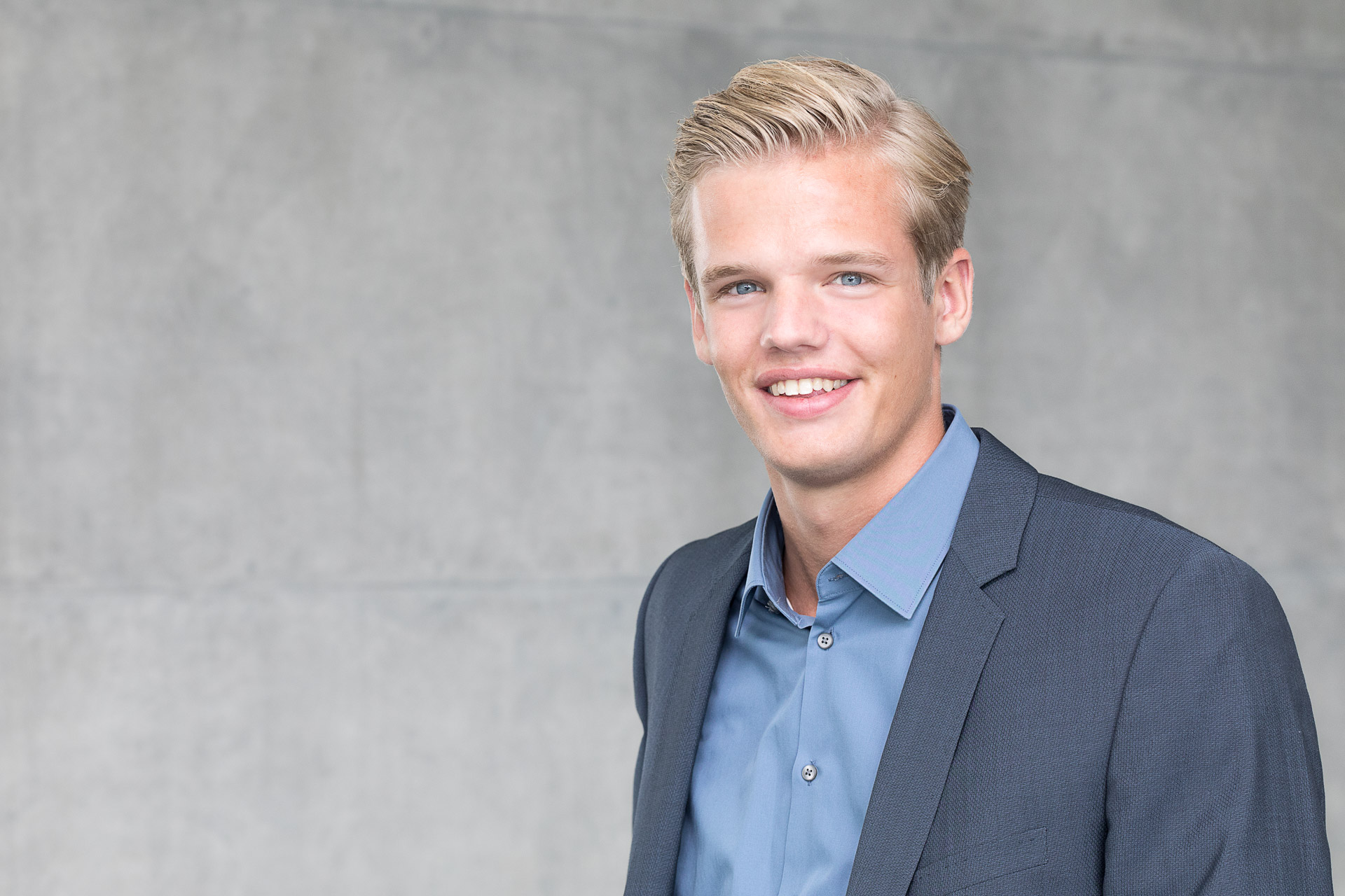 Portraitfotografie - Business Portraits: Lächelnder, junger Mann im blauen Anzug stehend vor grauer Wand
