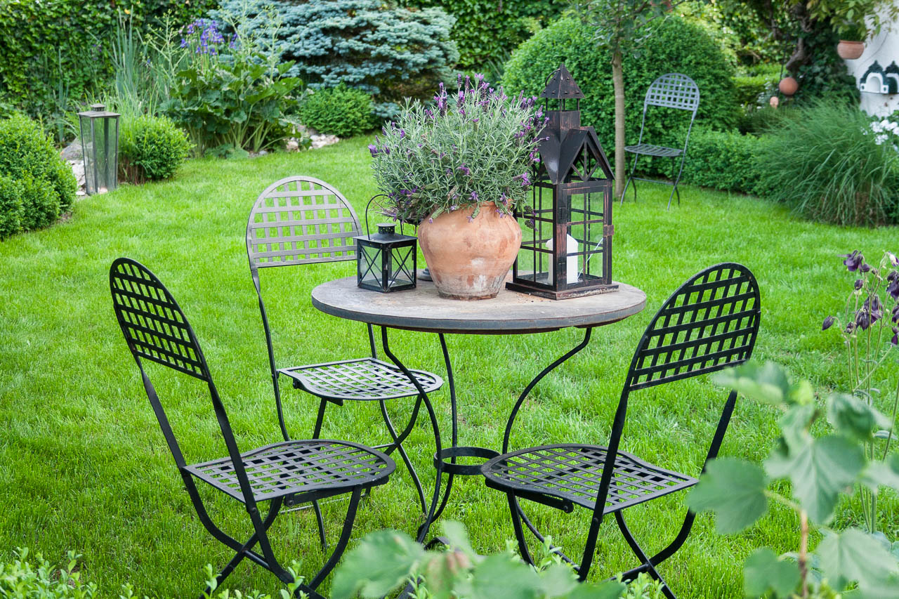 Gartenbilder: Sichtplatz im Rasen mit drei Metallstühlen. Auf dem Tisch zwei Laternen und eine Terrakotta Vase mit Lavendel bepflanzt